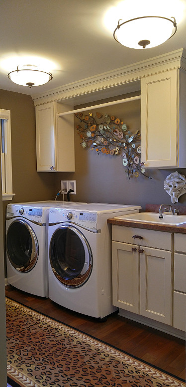 Photo of a country single-wall dedicated laundry room in New York with a drop-in sink, recessed-panel cabinets, white cabinets, laminate benchtops, brown walls, medium hardwood floors and a side-by-side washer and dryer.