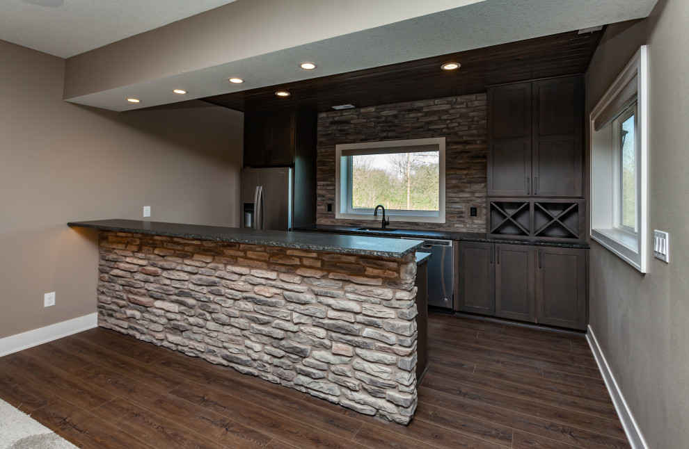 Photo of a transitional galley wet bar in Other with an undermount sink, shaker cabinets, dark wood cabinets, granite benchtops, stone slab splashback, vinyl floors, brown floor and black benchtop.