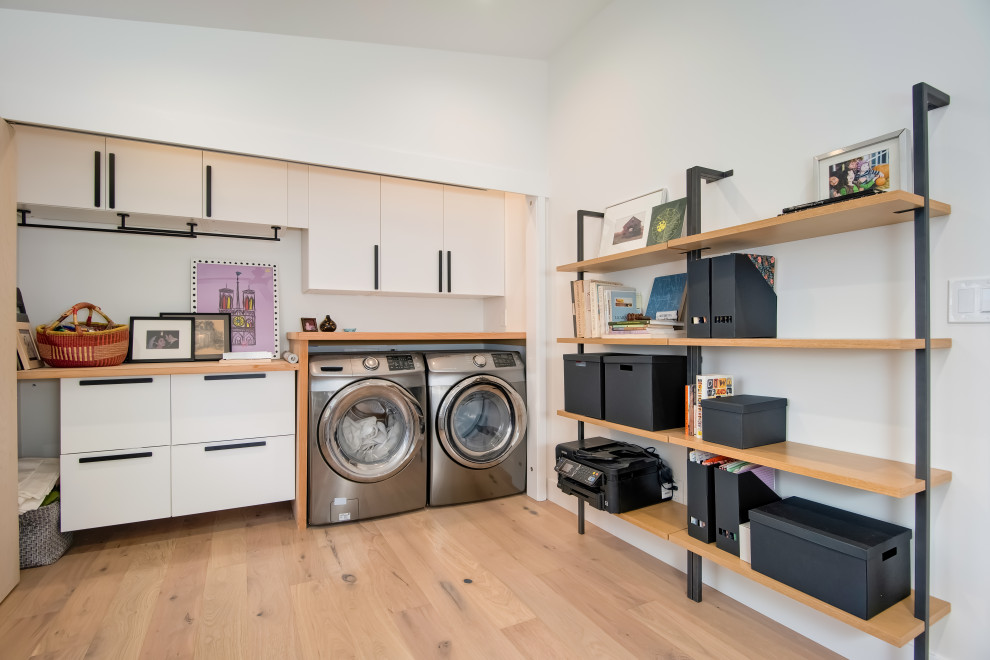 Photo of a mid-sized midcentury galley laundry cupboard in Orange County with flat-panel cabinets, white cabinets, wood benchtops, light hardwood floors and a concealed washer and dryer.