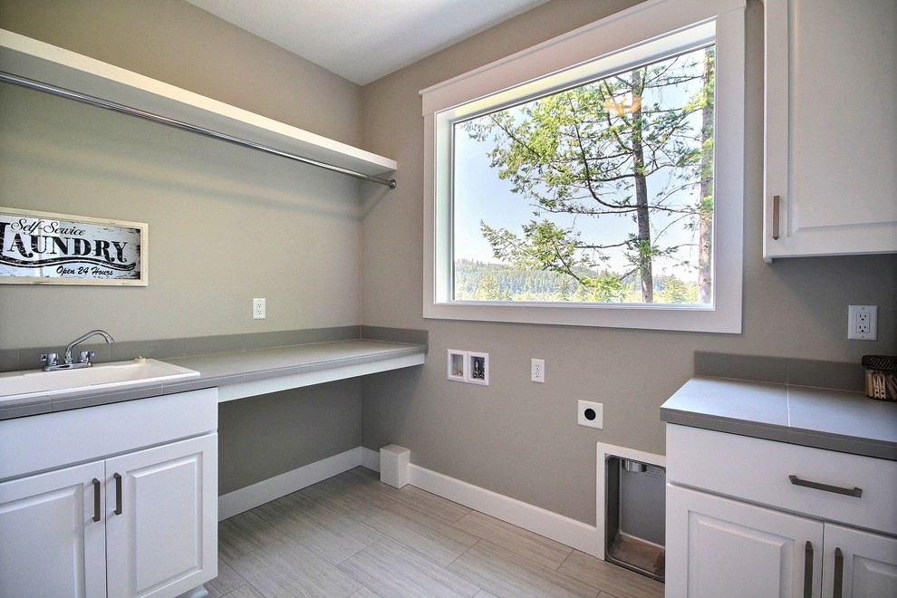 Large contemporary u-shaped utility room in Portland with a drop-in sink, raised-panel cabinets, white cabinets, tile benchtops, beige walls, porcelain floors, a side-by-side washer and dryer and grey floor.