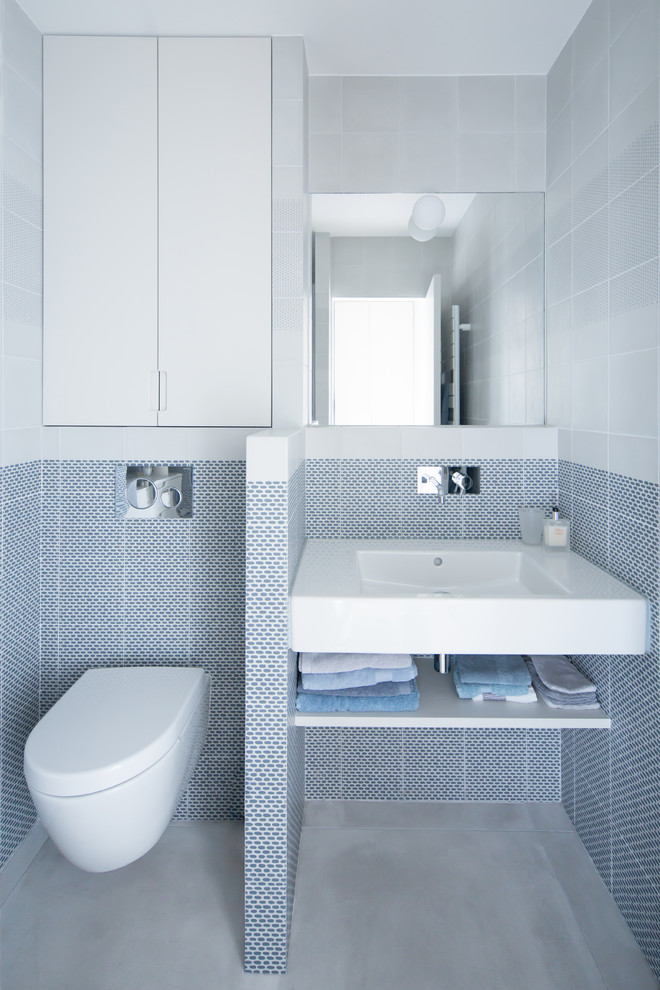 Photo of a small scandinavian 3/4 bathroom in Paris with open cabinets, blue tile, gray tile, concrete floors, a wall-mount sink, grey floor, a wall-mount toilet, white cabinets and white walls.