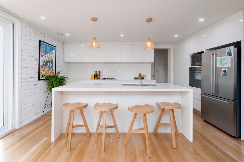 Photo of a mid-sized contemporary galley open plan kitchen in Canberra - Queanbeyan with white cabinets, white splashback, stainless steel appliances, light hardwood floors, with island, a double-bowl sink and flat-panel cabinets.