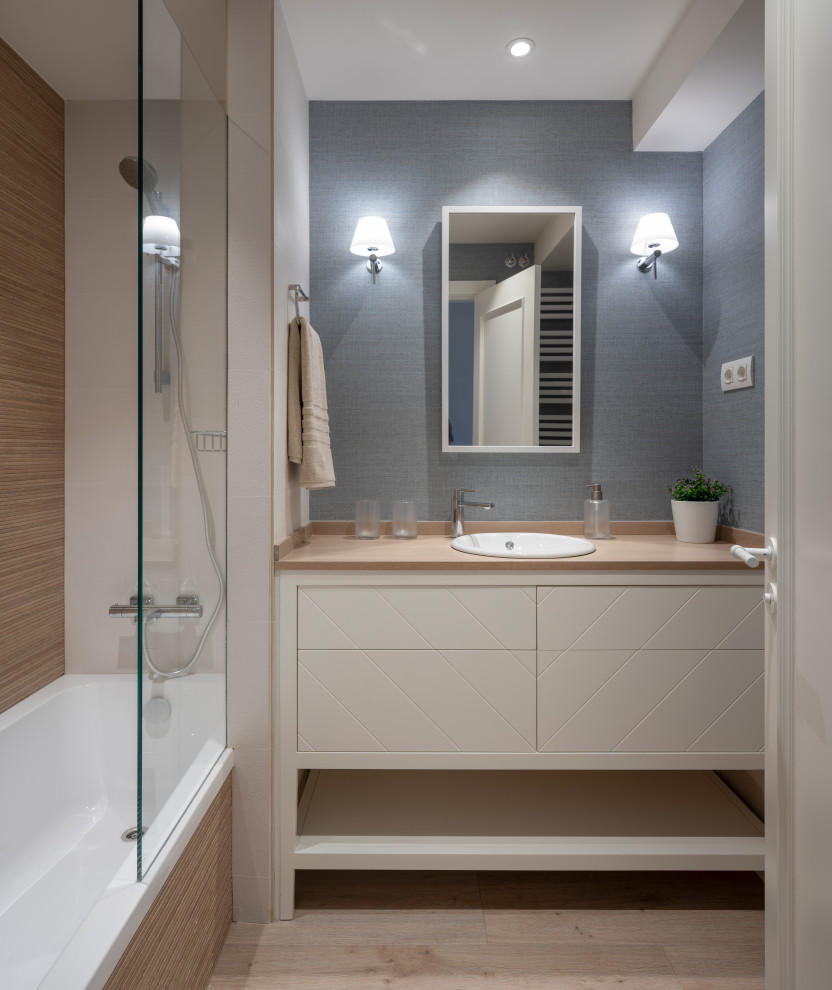 Example of a mid-sized transitional brown tile laminate floor, beige floor, single-sink and wallpaper alcove bathtub design in Bilbao with furniture-like cabinets, white cabinets, a wall-mount toilet, blue walls, a drop-in sink, wood countertops and brown countertops