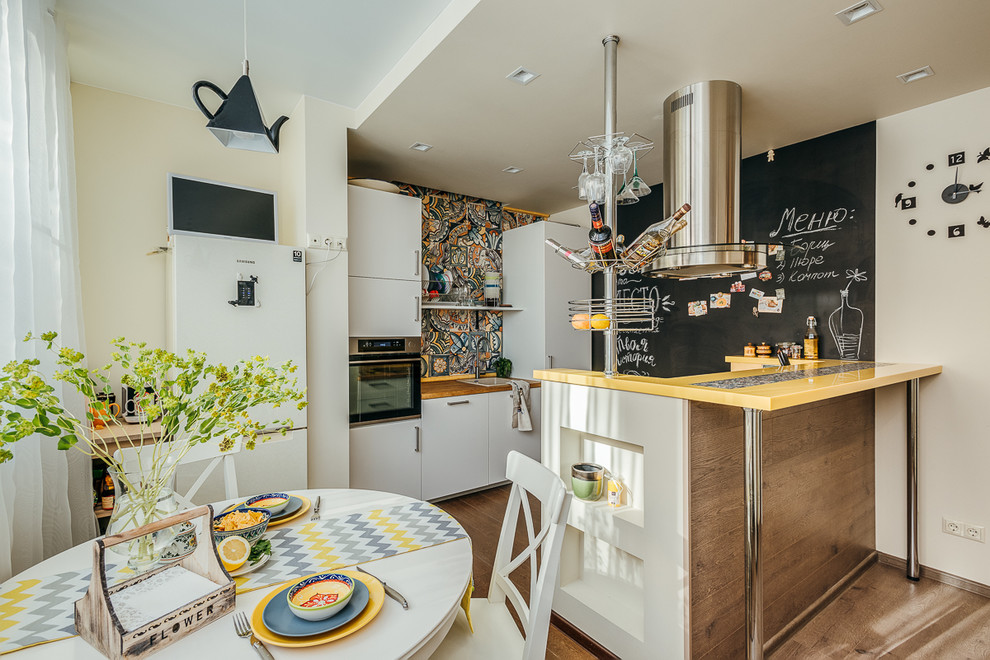 This is an example of a small eclectic galley eat-in kitchen in Moscow with a drop-in sink, flat-panel cabinets, white cabinets, multi-coloured splashback, stainless steel appliances, medium hardwood floors, with island, brown floor, yellow benchtop and ceramic splashback.