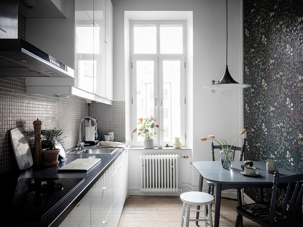 Inspiration for a small scandinavian eat-in kitchen in Gothenburg with flat-panel cabinets, no island, a drop-in sink, white cabinets, grey splashback, mosaic tile splashback, light hardwood floors, beige floor and black benchtop.
