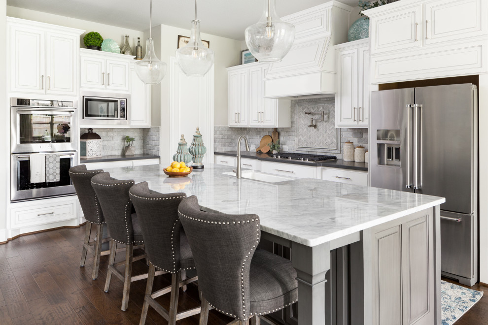 This is an example of a traditional l-shaped kitchen in Houston with a farmhouse sink, raised-panel cabinets, white cabinets, grey splashback, stainless steel appliances, dark hardwood floors, with island, brown floor and white benchtop.