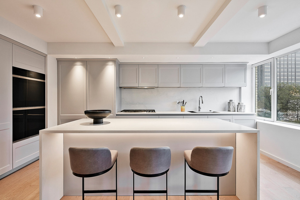 Photo of a mid-sized transitional l-shaped eat-in kitchen in New York with a double-bowl sink, recessed-panel cabinets, grey cabinets, quartz benchtops, white splashback, stone slab splashback, stainless steel appliances, medium hardwood floors, with island, yellow floor and white benchtop.