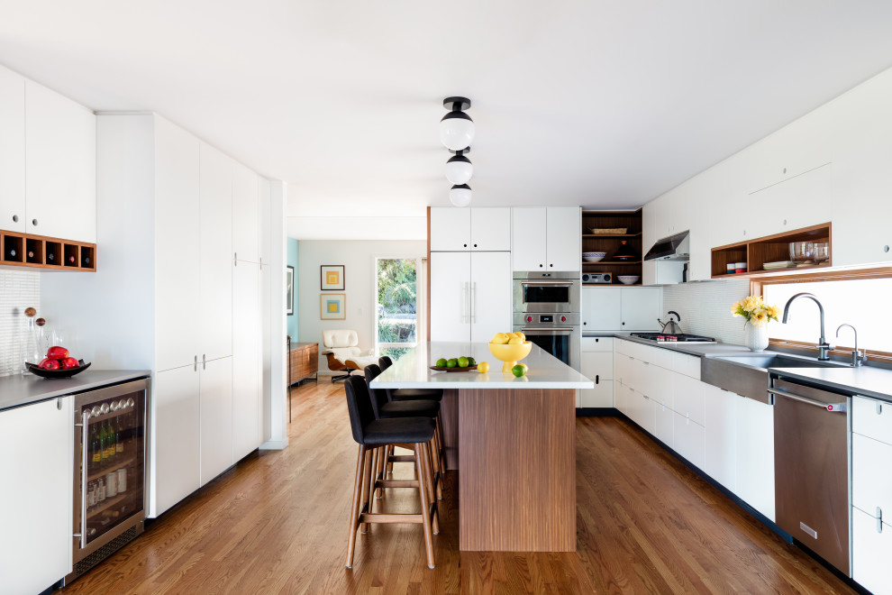 Medium sized midcentury l-shaped kitchen/diner in Seattle with flat-panel cabinets, white cabinets, composite countertops, stainless steel appliances, medium hardwood flooring, an island, white worktops, a belfast sink, white splashback, mosaic tiled splashback and brown floors.