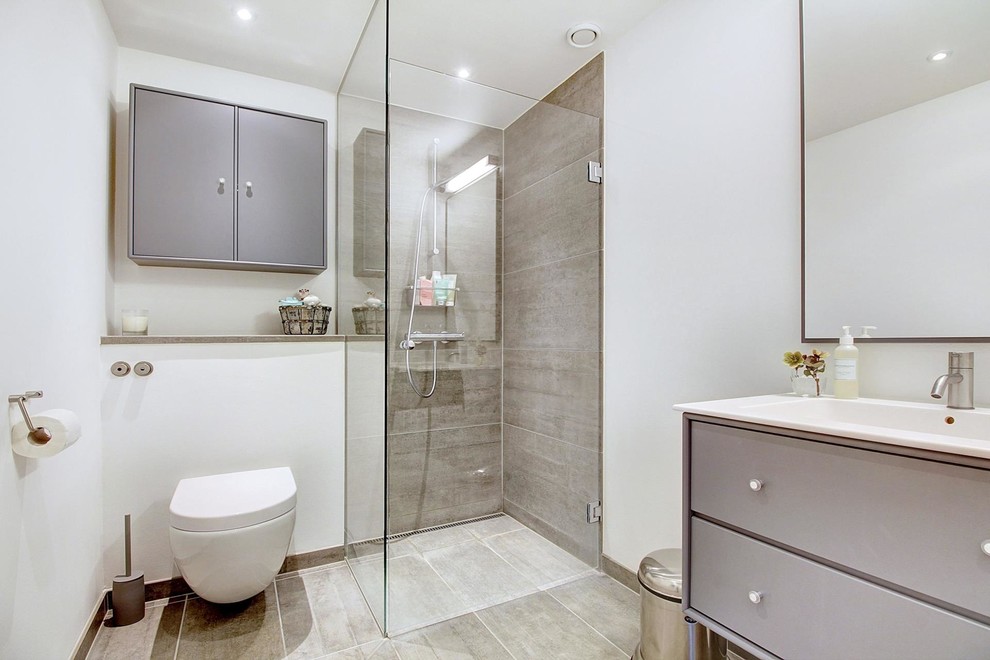 Transitional bathroom in Copenhagen with flat-panel cabinets, grey cabinets, a corner shower, a wall-mount toilet, gray tile, white walls, ceramic floors, an integrated sink and a hinged shower door.