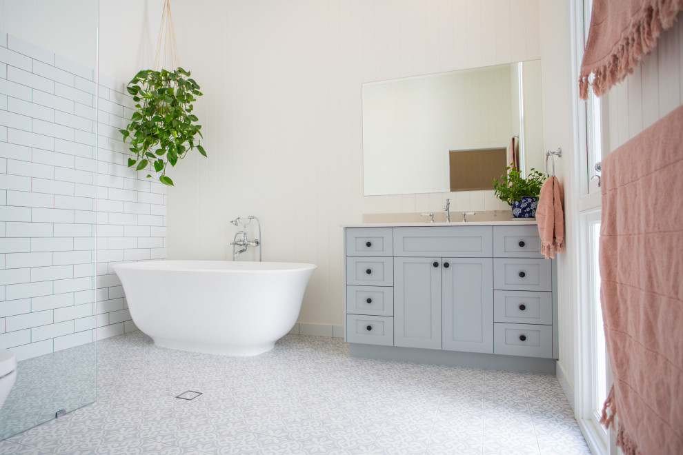 This is an example of a large contemporary master bathroom in Brisbane with furniture-like cabinets, grey cabinets, a freestanding tub, a corner shower, a one-piece toilet, white tile, white walls, ceramic floors, a drop-in sink, marble benchtops, blue floor and white benchtops.
