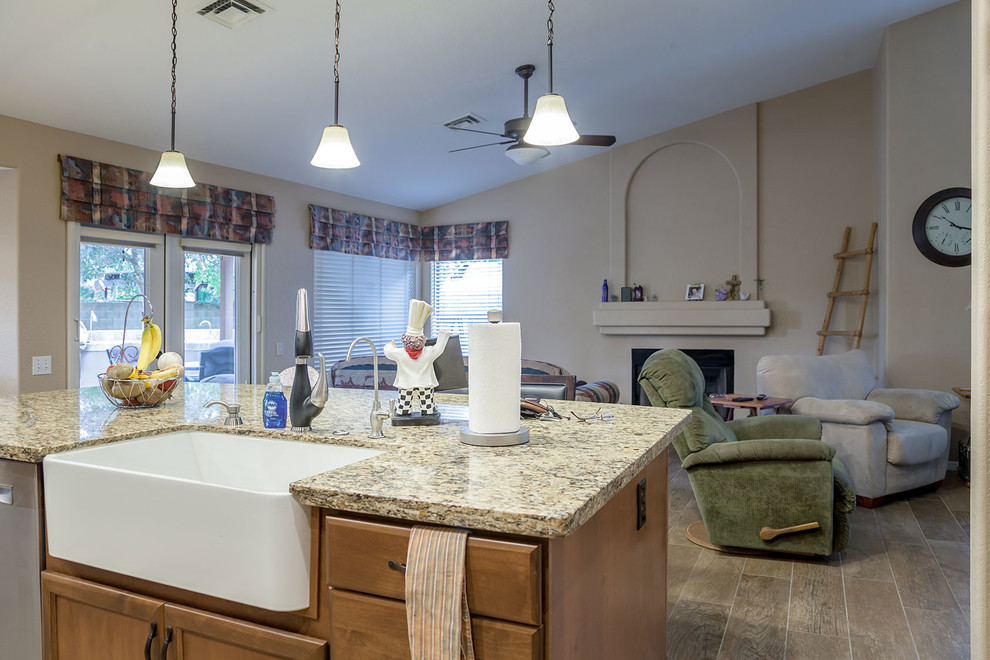 Mid-sized transitional l-shaped laminate floor and brown floor open concept kitchen photo in Phoenix with a farmhouse sink, recessed-panel cabinets, medium tone wood cabinets, granite countertops, beige backsplash, stone slab backsplash, stainless steel appliances and an island