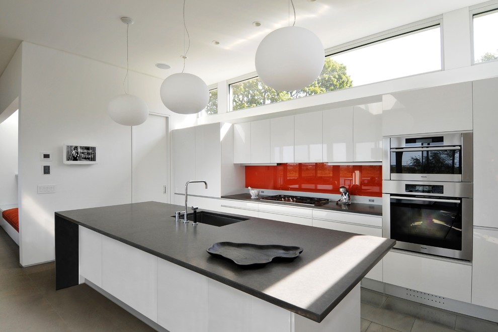Photo of a large contemporary galley eat-in kitchen in New York with an undermount sink, flat-panel cabinets, white cabinets, red splashback, glass sheet splashback, stainless steel appliances, with island, quartz benchtops and porcelain floors.