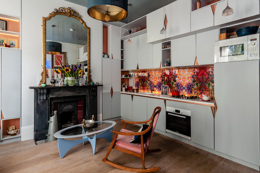 Photo of a small eclectic single-wall open plan kitchen in London with an integrated sink, flat-panel cabinets, grey cabinets, solid surface benchtops, multi-coloured splashback, glass sheet splashback, medium hardwood floors, no island, brown floor, white benchtop and white appliances.