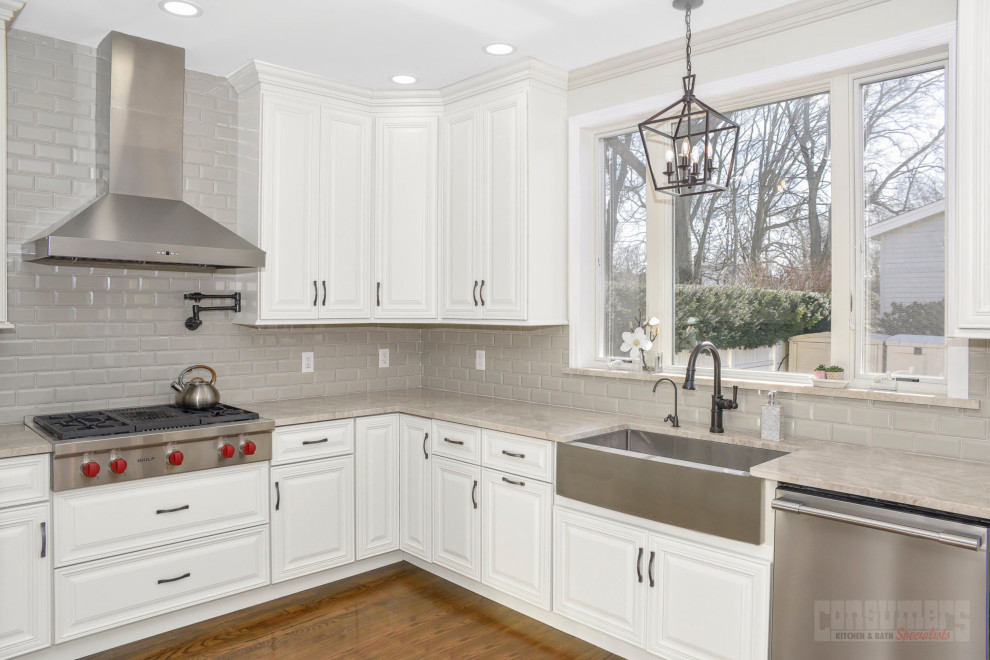 This is an example of a mid-sized transitional u-shaped eat-in kitchen in New York with a farmhouse sink, raised-panel cabinets, white cabinets, quartzite benchtops, grey splashback, ceramic splashback, stainless steel appliances, medium hardwood floors, with island, brown floor and beige benchtop.