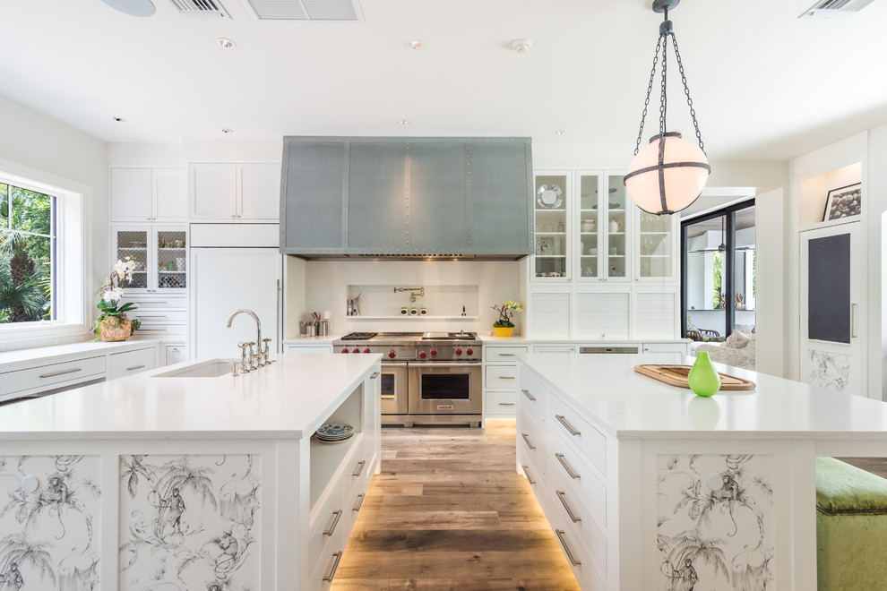 Photo of a tropical u-shaped kitchen in Miami with granite benchtops, white splashback, stainless steel appliances, medium hardwood floors, an undermount sink, multiple islands and brown floor.