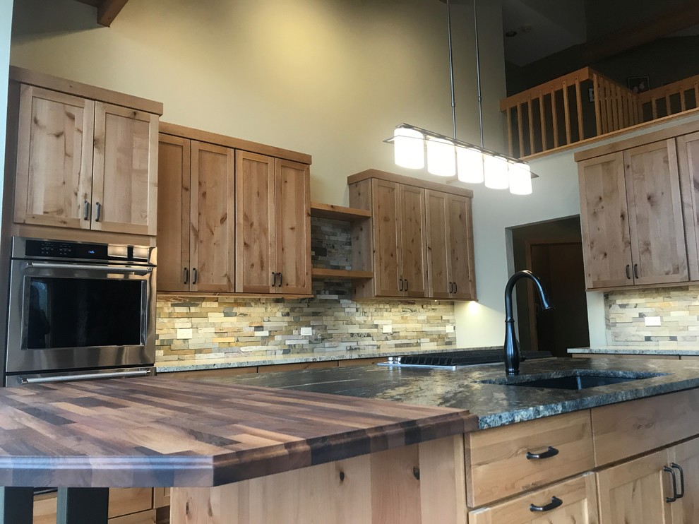 This is an example of a large country l-shaped open plan kitchen in Chicago with an undermount sink, shaker cabinets, medium wood cabinets, granite benchtops, green splashback, slate splashback, stainless steel appliances, medium hardwood floors, with island, brown floor and black benchtop.