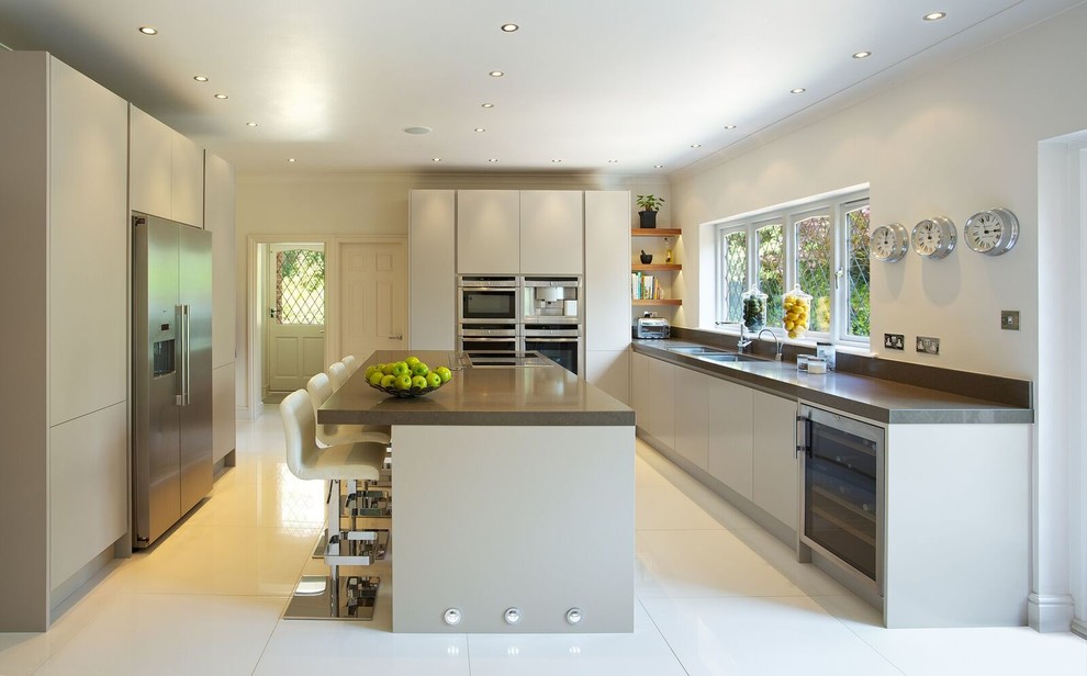 This is an example of a contemporary galley eat-in kitchen in Hertfordshire with a double-bowl sink, flat-panel cabinets, grey cabinets, stainless steel appliances, with island, solid surface benchtops and porcelain floors.