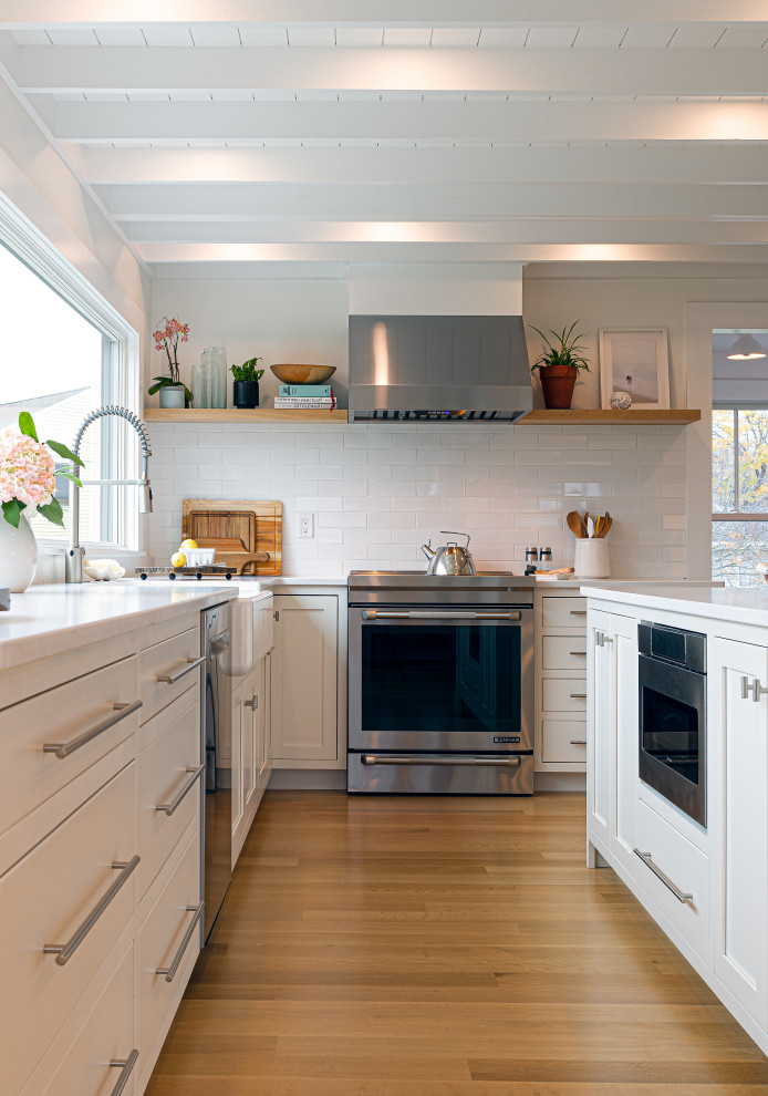 Contemporary l-shaped eat-in kitchen in Boston with a farmhouse sink, shaker cabinets, white cabinets, quartz benchtops, white splashback, porcelain splashback, stainless steel appliances, with island and white benchtop.