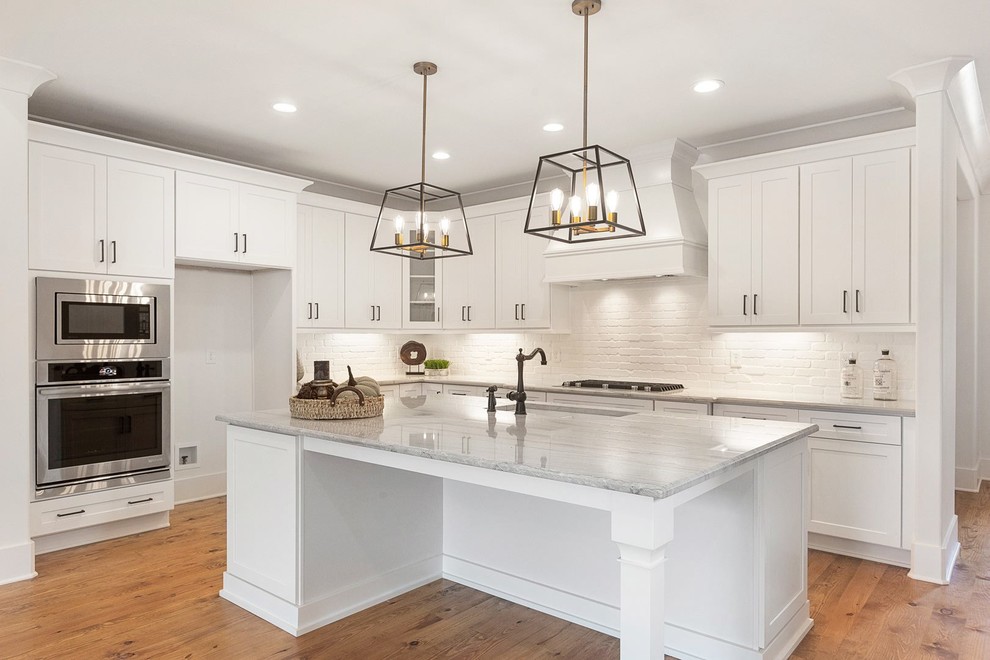 Photo of a mid-sized country l-shaped eat-in kitchen in Raleigh with an undermount sink, shaker cabinets, white cabinets, granite benchtops, white splashback, brick splashback, stainless steel appliances, medium hardwood floors, with island and white benchtop.