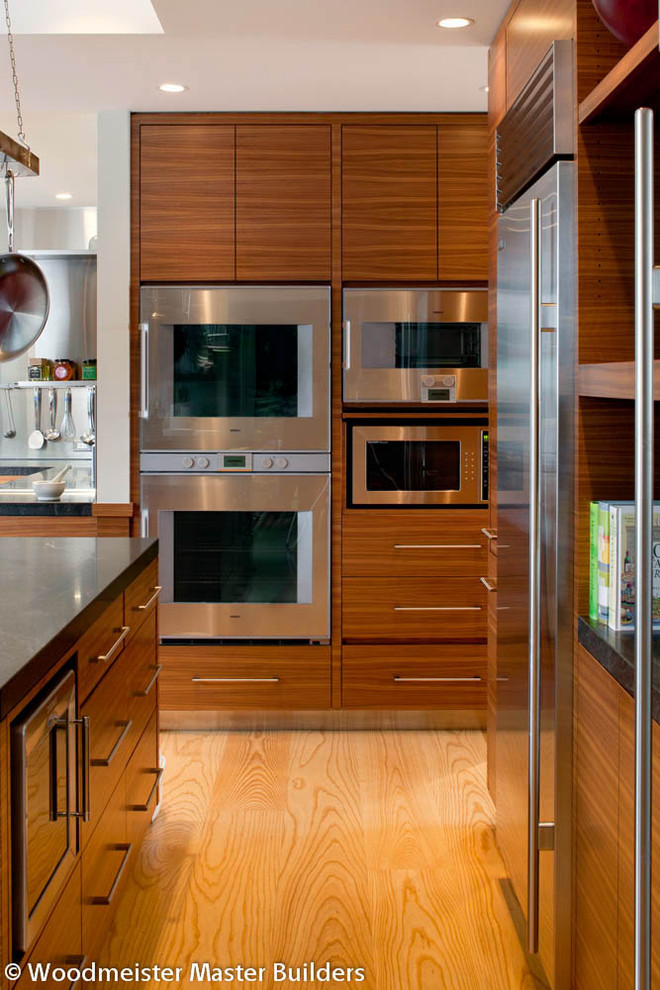 This is an example of a mid-sized contemporary galley kitchen in Boston with flat-panel cabinets, medium wood cabinets, granite benchtops, stainless steel appliances, light hardwood floors and with island.