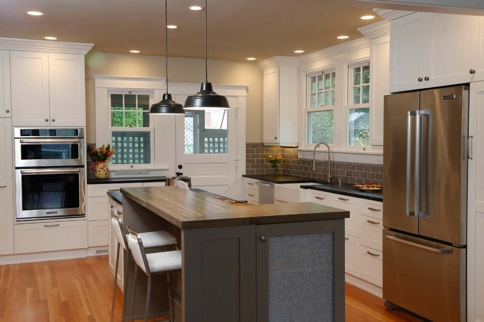 Inspiration for a mid-sized transitional l-shaped medium tone wood floor eat-in kitchen remodel in Seattle with a farmhouse sink, shaker cabinets, white cabinets, soapstone countertops, brown backsplash, ceramic backsplash, stainless steel appliances and an island