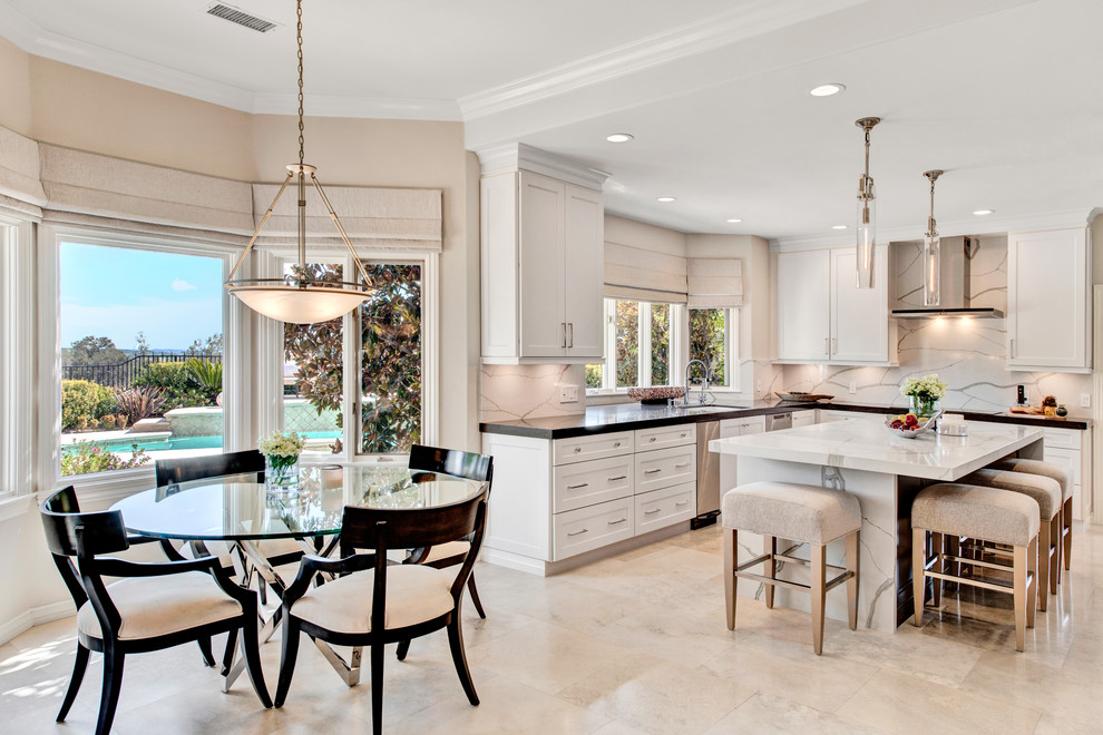 This is an example of a transitional u-shaped eat-in kitchen in Los Angeles with an undermount sink, shaker cabinets, white cabinets, white splashback, stone slab splashback, with island, beige floor and black benchtop.