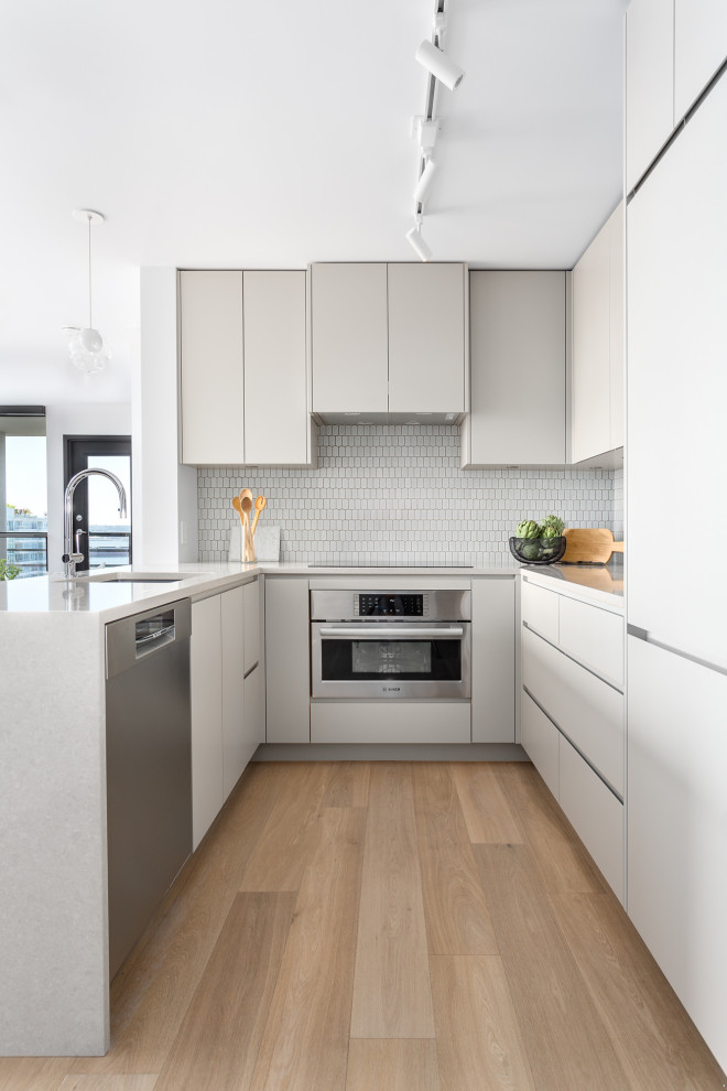 Example of a small trendy u-shaped light wood floor and brown floor eat-in kitchen design in Vancouver with an undermount sink, flat-panel cabinets, beige cabinets, quartzite countertops, white backsplash, mosaic tile backsplash, stainless steel appliances, a peninsula and white countertops
