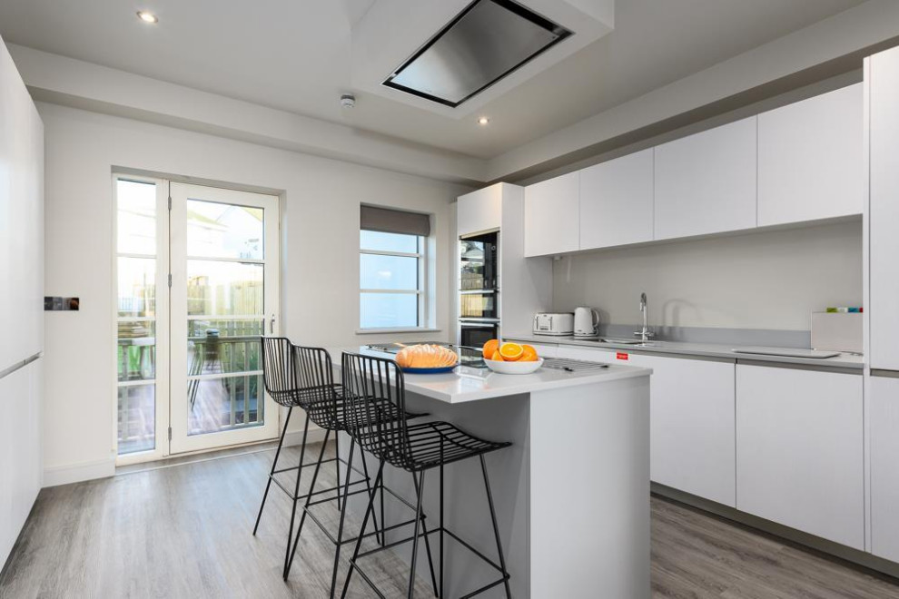 Photo of a mid-sized beach style galley eat-in kitchen in Cornwall with a drop-in sink, flat-panel cabinets, white cabinets, white splashback, engineered quartz splashback, black appliances, light hardwood floors, with island, grey floor, white benchtop and coffered.