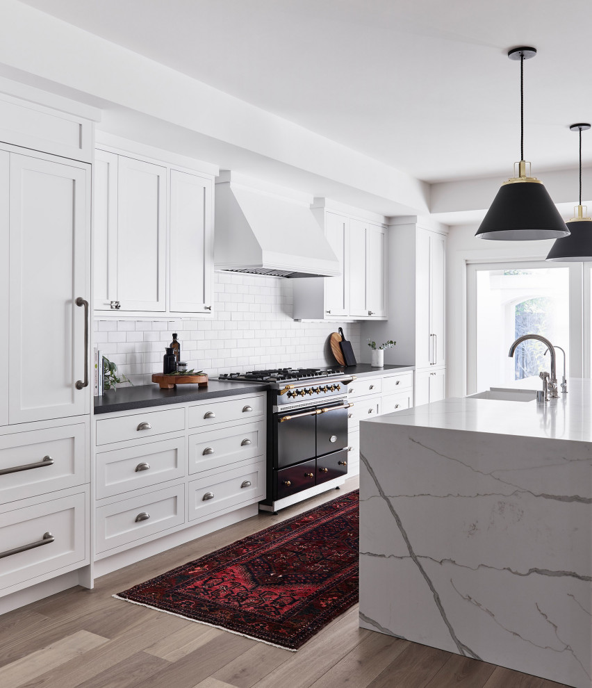 Inspiration for a large transitional galley kitchen in Chicago with a farmhouse sink, shaker cabinets, white cabinets, white splashback, subway tile splashback, panelled appliances, with island, beige floor and black benchtop.