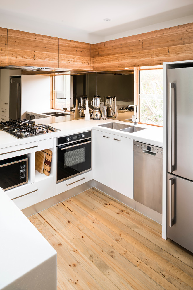 This is an example of a small contemporary u-shaped eat-in kitchen in Melbourne with a double-bowl sink, white cabinets, solid surface benchtops, mirror splashback, stainless steel appliances, no island, flat-panel cabinets and light hardwood floors.