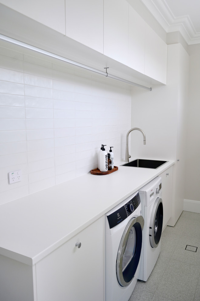 This is an example of a mid-sized transitional single-wall dedicated laundry room in Sydney with a drop-in sink, flat-panel cabinets, white cabinets, laminate benchtops, white splashback, subway tile splashback, white walls, concrete floors, a side-by-side washer and dryer, grey floor and white benchtop.