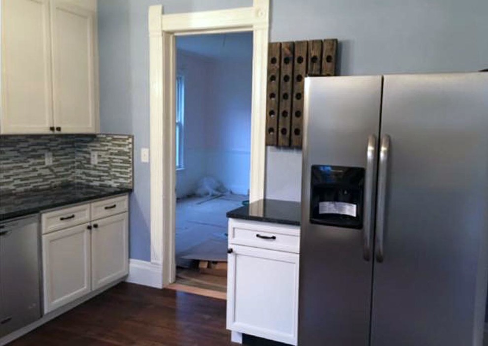 Example of a mid-sized transitional l-shaped medium tone wood floor and brown floor kitchen pantry design with a farmhouse sink, recessed-panel cabinets, white cabinets, granite countertops, multicolored backsplash, glass tile backsplash, stainless steel appliances, no island and black countertops