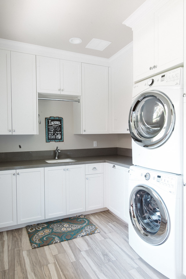 This is an example of a large country l-shaped dedicated laundry room in Boston with an undermount sink, white cabinets, solid surface benchtops, grey walls, a stacked washer and dryer, shaker cabinets and plywood floors.