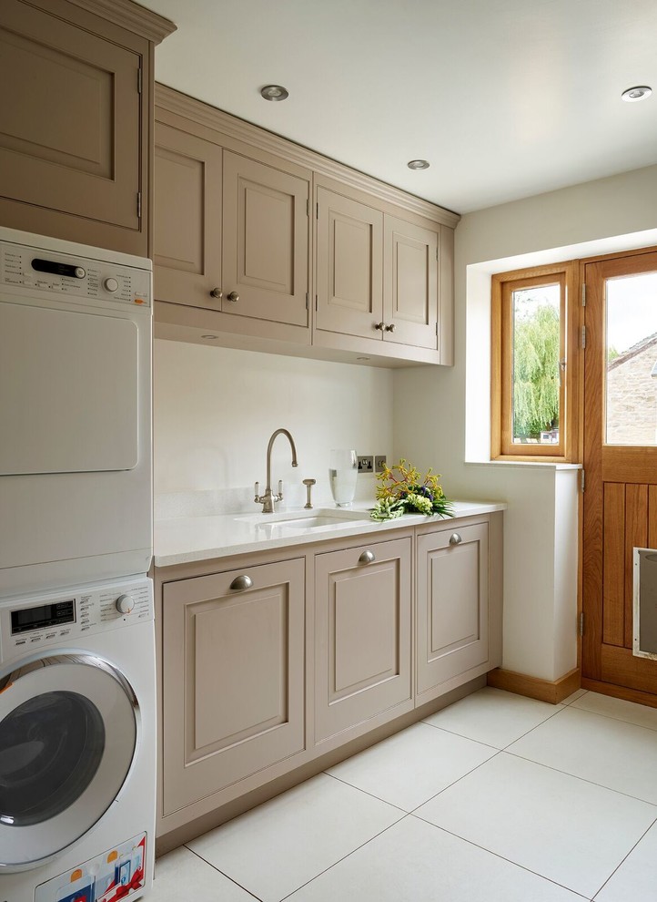 Inspiration for a mid-sized traditional single-wall utility room in Other with an undermount sink, recessed-panel cabinets, brown cabinets, beige walls, a stacked washer and dryer and white floor.