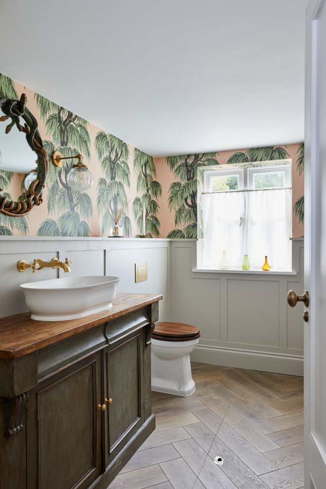 Klassisk inredning av ett brun brunt toalett, med luckor med infälld panel, skåp i mellenmörkt trä, en toalettstol med separat cisternkåpa, flerfärgade väggar, mellanmörkt trägolv, ett fristående handfat, träbänkskiva och brunt golv