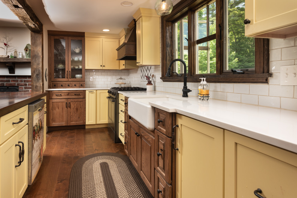 Design ideas for a large country l-shaped kitchen in Portland Maine with a farmhouse sink, shaker cabinets, yellow cabinets, quartz benchtops, white splashback, subway tile splashback, stainless steel appliances, dark hardwood floors, with island, brown floor and white benchtop.