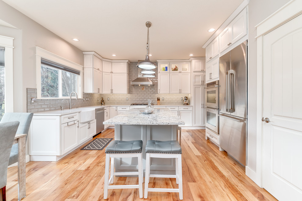 Example of a mid-sized transitional u-shaped medium tone wood floor and brown floor eat-in kitchen design in Boise with a farmhouse sink, shaker cabinets, white cabinets, quartz countertops, gray backsplash, subway tile backsplash, stainless steel appliances and an island