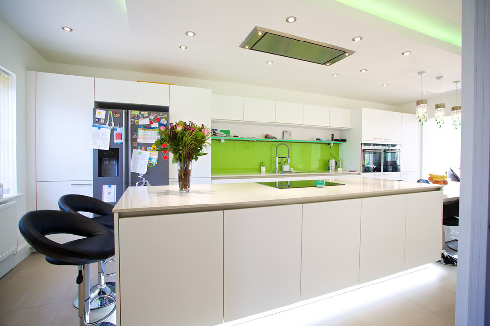 Design ideas for a mid-sized contemporary galley eat-in kitchen in Hampshire with flat-panel cabinets, white cabinets, green splashback, glass sheet splashback, stainless steel appliances, an integrated sink, solid surface benchtops, porcelain floors and with island.