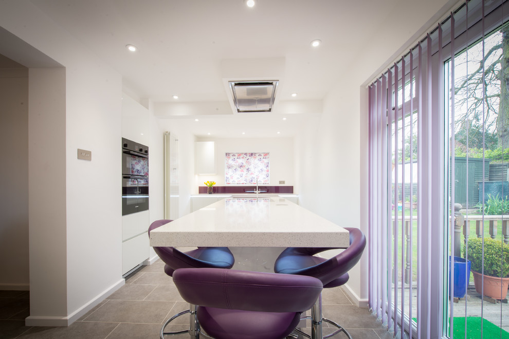 This is an example of a mid-sized contemporary open plan kitchen in Other with glass-front cabinets, white cabinets, solid surface benchtops, pink splashback, glass tile splashback, black appliances, ceramic floors, with island, grey floor and white benchtop.