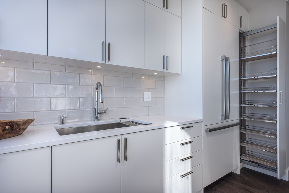 This is an example of a mid-sized contemporary galley kitchen in Vancouver with an undermount sink, flat-panel cabinets, white cabinets, quartz benchtops, grey splashback, porcelain splashback, panelled appliances, medium hardwood floors, a peninsula, brown floor and white benchtop.