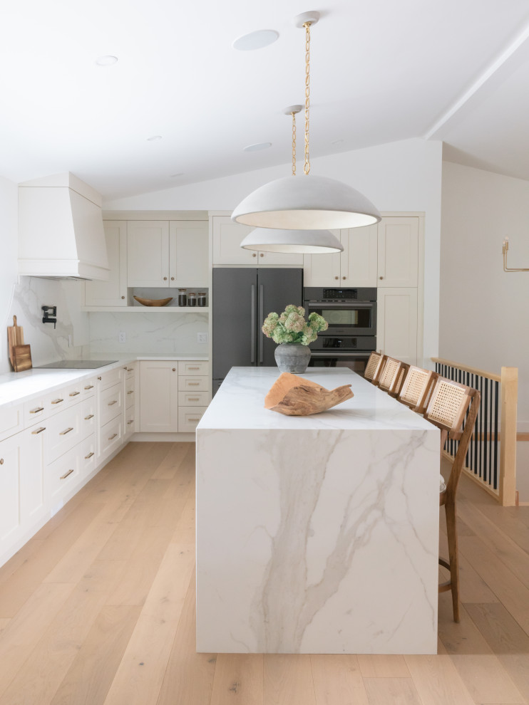 Example of a transitional l-shaped light wood floor, beige floor and vaulted ceiling kitchen design with shaker cabinets, beige cabinets, white backsplash, stone slab backsplash, stainless steel appliances, an island and white countertops