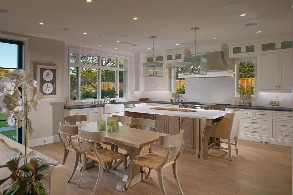 Inspiration for a beach style l-shaped eat-in kitchen in Orange County with a farmhouse sink, recessed-panel cabinets, white cabinets, white splashback, stone slab splashback and light hardwood floors.