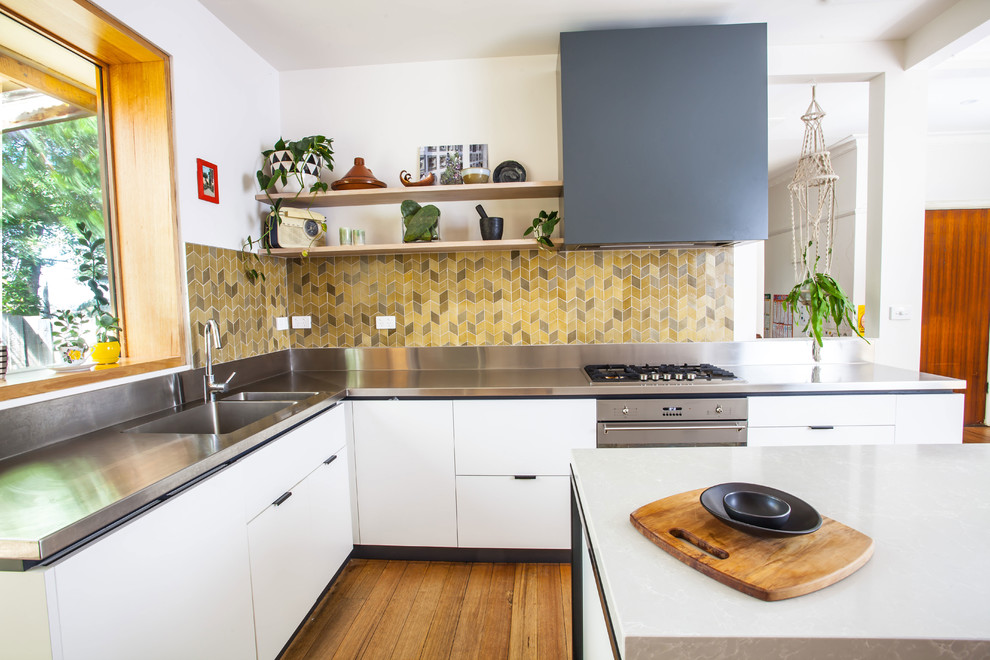 Mid-sized contemporary u-shaped open plan kitchen in Melbourne with a double-bowl sink, flat-panel cabinets, white cabinets, stainless steel benchtops, green splashback, ceramic splashback, stainless steel appliances, medium hardwood floors and with island.