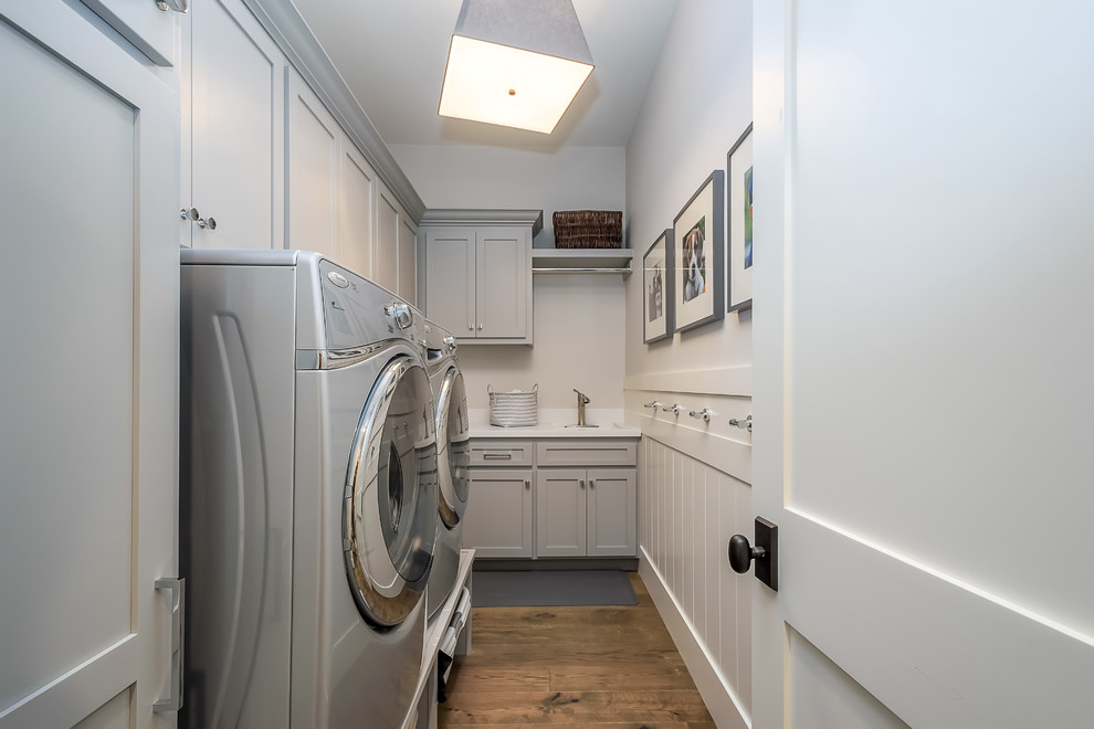 Photo of a mid-sized contemporary l-shaped utility room in San Francisco with an undermount sink, shaker cabinets, grey cabinets, solid surface benchtops, grey walls, medium hardwood floors, a side-by-side washer and dryer and brown floor.