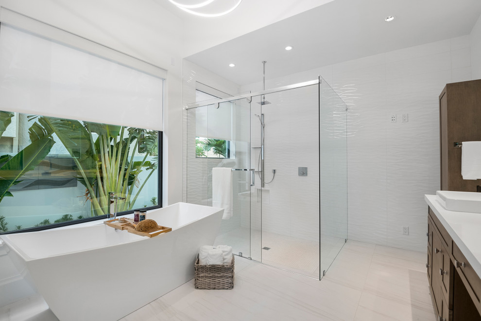 This is an example of a mid-sized contemporary master bathroom in Miami with flat-panel cabinets, light wood cabinets, a freestanding tub, an open shower, a one-piece toilet, gray tile, porcelain tile, white walls, porcelain floors, a vessel sink, engineered quartz benchtops, white floor, a sliding shower screen, white benchtops, an enclosed toilet, a double vanity, a freestanding vanity and recessed.
