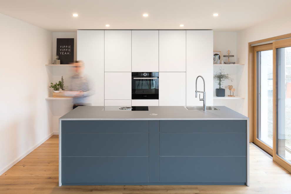 This is an example of a large modern single-wall separate kitchen in Stuttgart with a single-bowl sink, flat-panel cabinets, white cabinets, laminate benchtops, white splashback, timber splashback, black appliances, with island, black benchtop, light hardwood floors and brown floor.