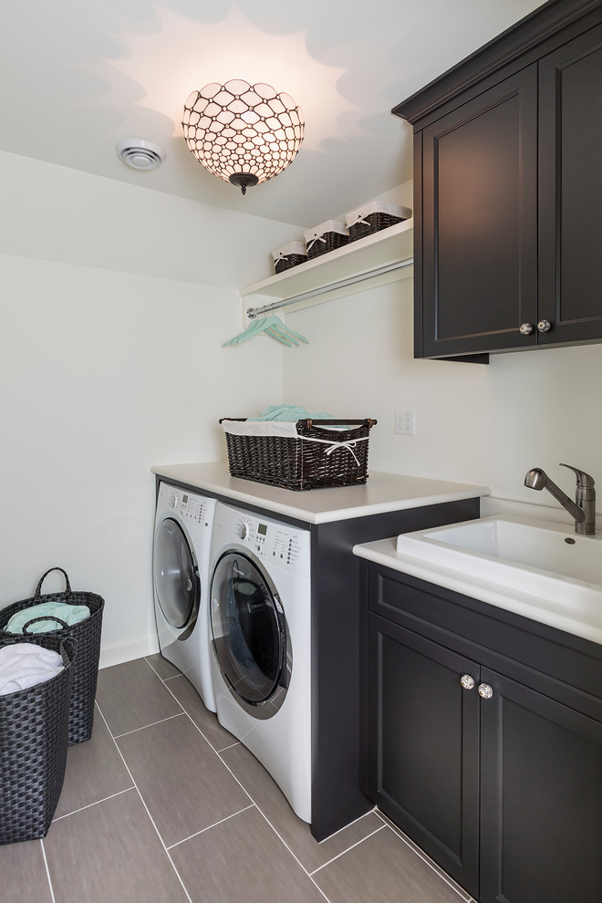 Photo of a mid-sized transitional single-wall dedicated laundry room in Minneapolis with a drop-in sink, black cabinets, laminate benchtops, white walls, porcelain floors, a side-by-side washer and dryer and shaker cabinets.