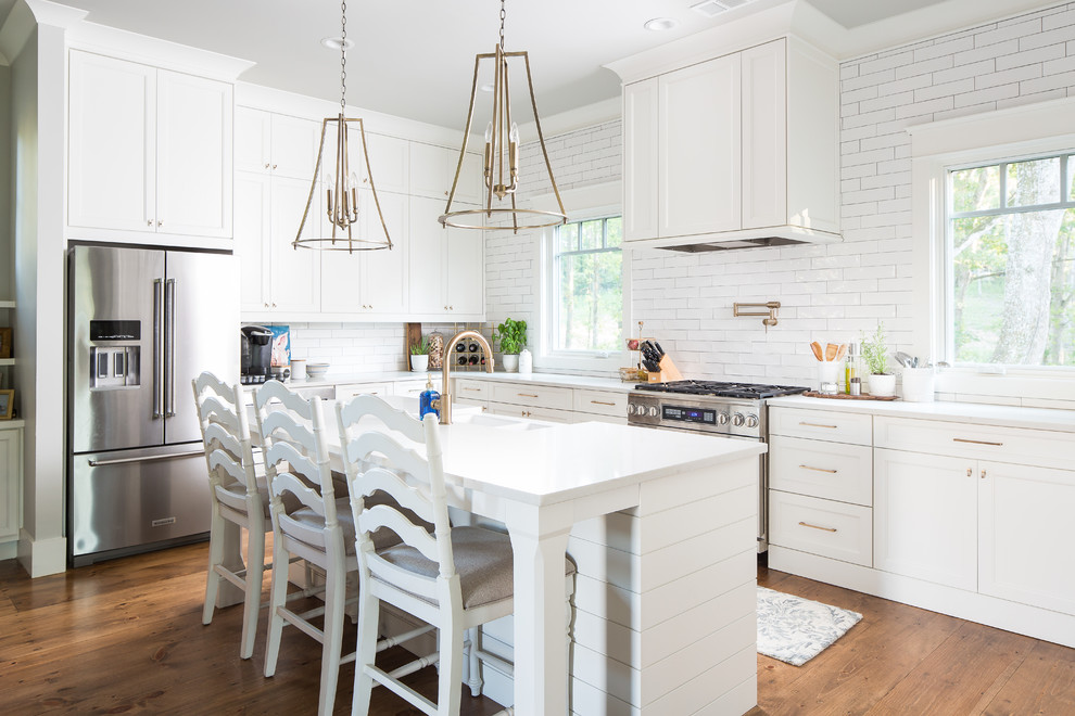 Inspiration for a country l-shaped kitchen in Atlanta with a farmhouse sink, recessed-panel cabinets, white cabinets, white splashback, subway tile splashback, stainless steel appliances, medium hardwood floors, with island, brown floor and white benchtop.