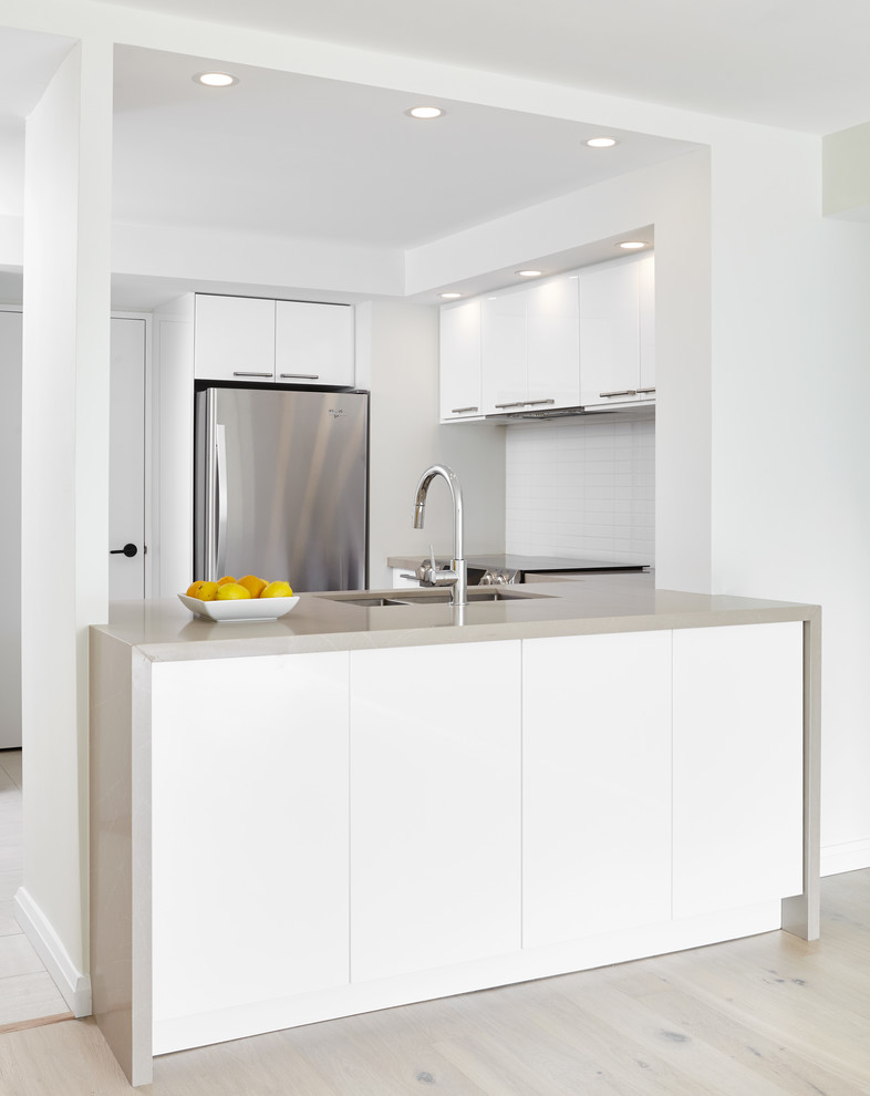 Photo of a mid-sized modern u-shaped separate kitchen in Toronto with a double-bowl sink, flat-panel cabinets, white cabinets, solid surface benchtops, white splashback, ceramic splashback, stainless steel appliances, light hardwood floors, a peninsula and brown floor.