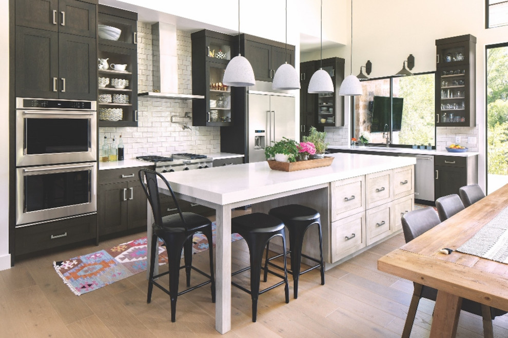Design ideas for a mid-sized country l-shaped eat-in kitchen in Denver with recessed-panel cabinets, quartz benchtops, white splashback, ceramic splashback, stainless steel appliances, medium hardwood floors, with island, brown floor, white benchtop, black cabinets and an undermount sink.